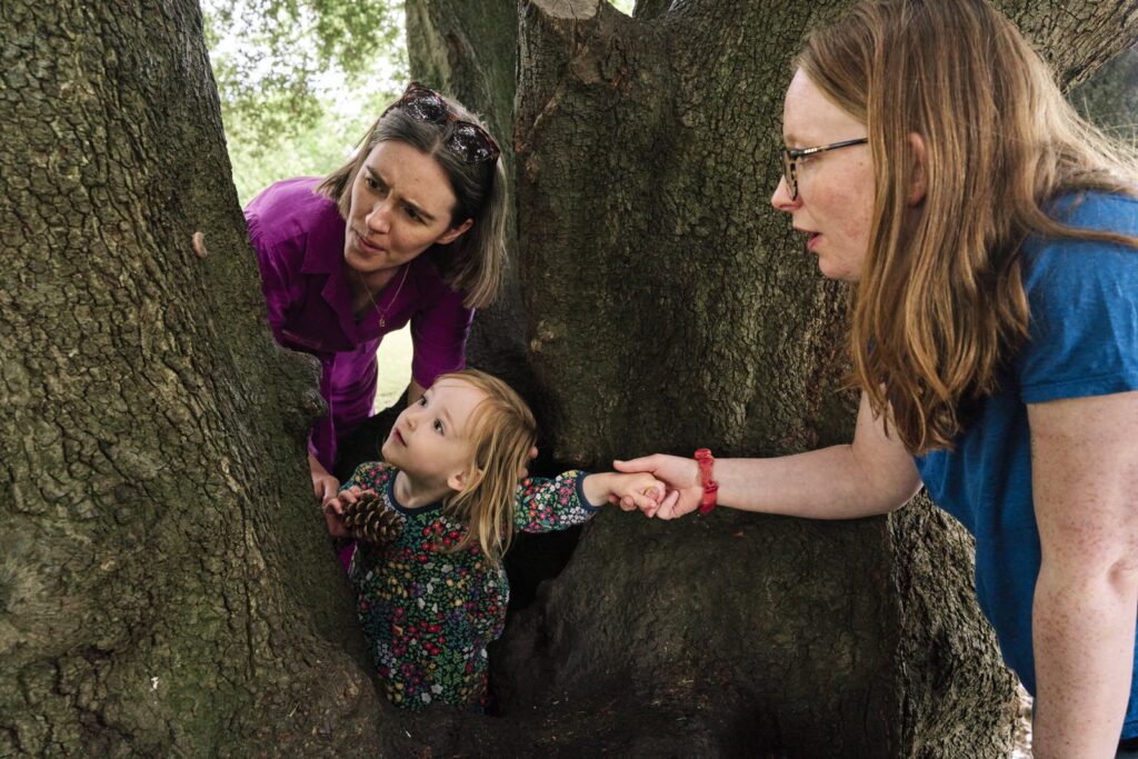 Child climbing a tree with help from mum to see a large caterpillar during a storytelling family photo session in a park in London