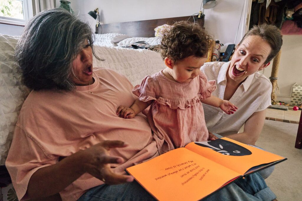 Documentary-style unposed family photo of two mums reading a book to her child on the floor in a London home